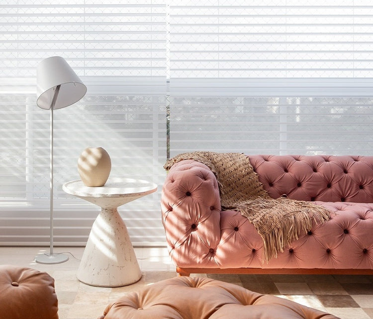 mesa lateral com base e tampo cônico que se unem ao lado de um sofá chesterfiel em tecido rosa ambiente claro a frente de uma grande janela com persianas brancas