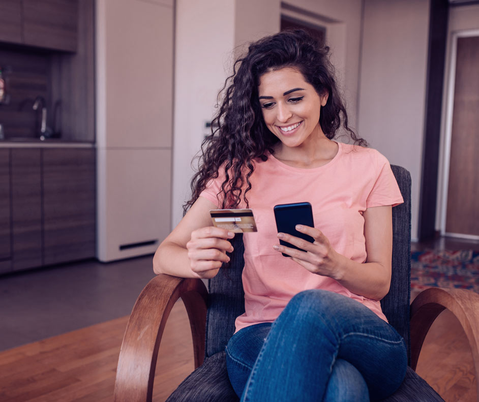 garota fazendo uma compra pelo celular sentada em sua poltronas na sua casa, em uma das mão segura cartão de crédito na outra o celular