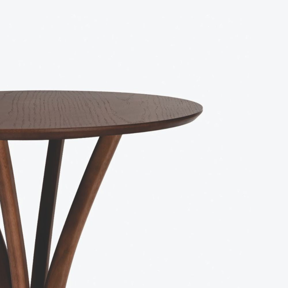 Detalhe lateral e aproximado da mesa bistrô montana, com tampo redondo com bordas chanfradas e pés em madeira escultural curvas.