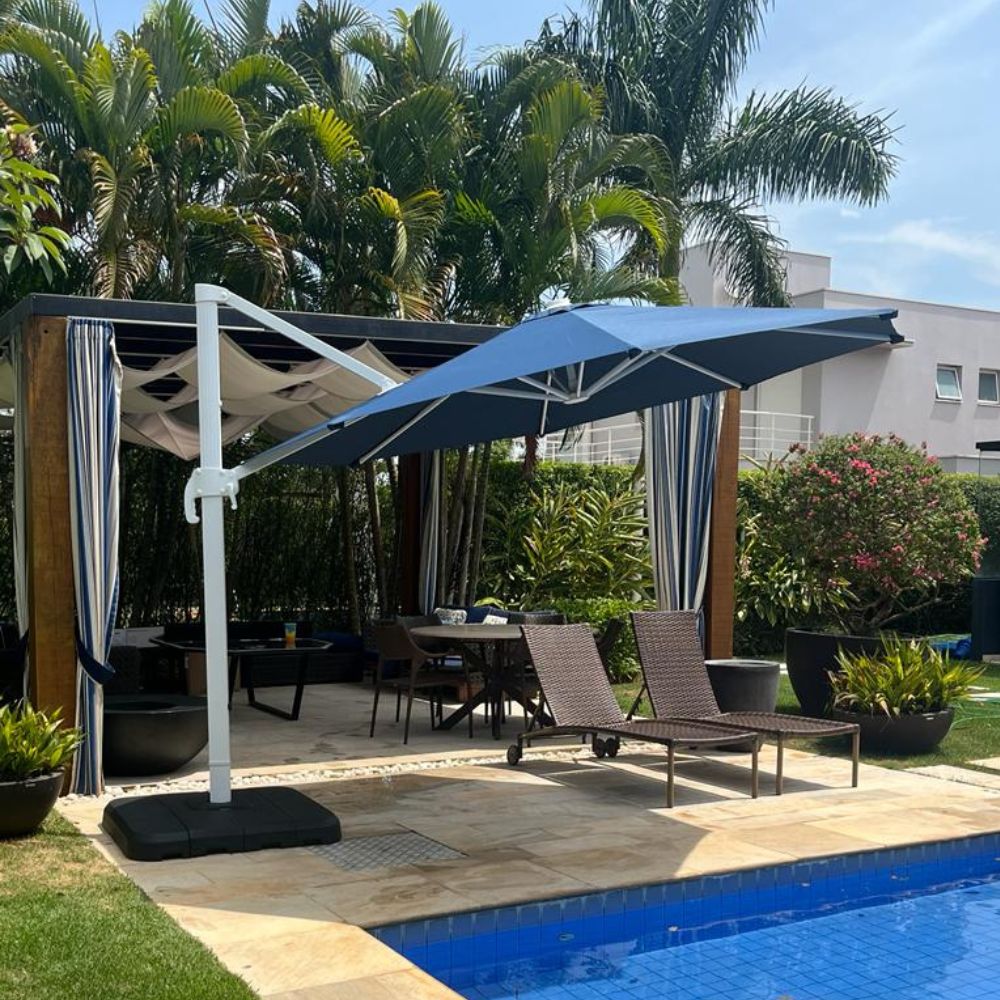 Ombrelone Cantilever azul marinho com mastro lateral, sombreando área de estar e espreguiçadeiras de vime à beira da piscina.