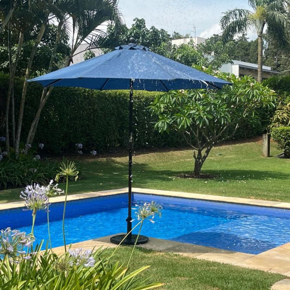 Ombrelone Santorini de centro com cobertura branca/cinza claro, protegendo área de estar externa com mesa e cadeiras em um jardim.