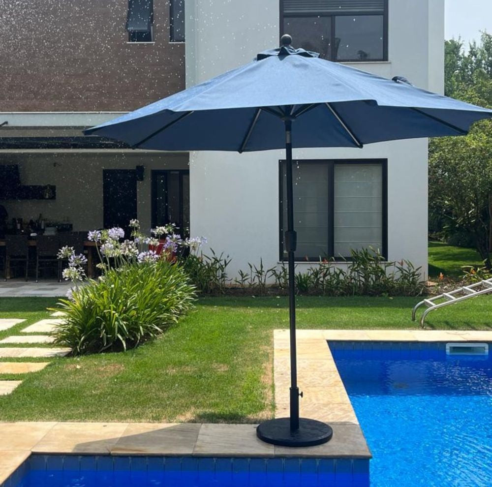 Ombrelone Santorini de centro, com cobertura em tecido azul marinho, sombreando a borda de uma piscina em área de lazer residencial.