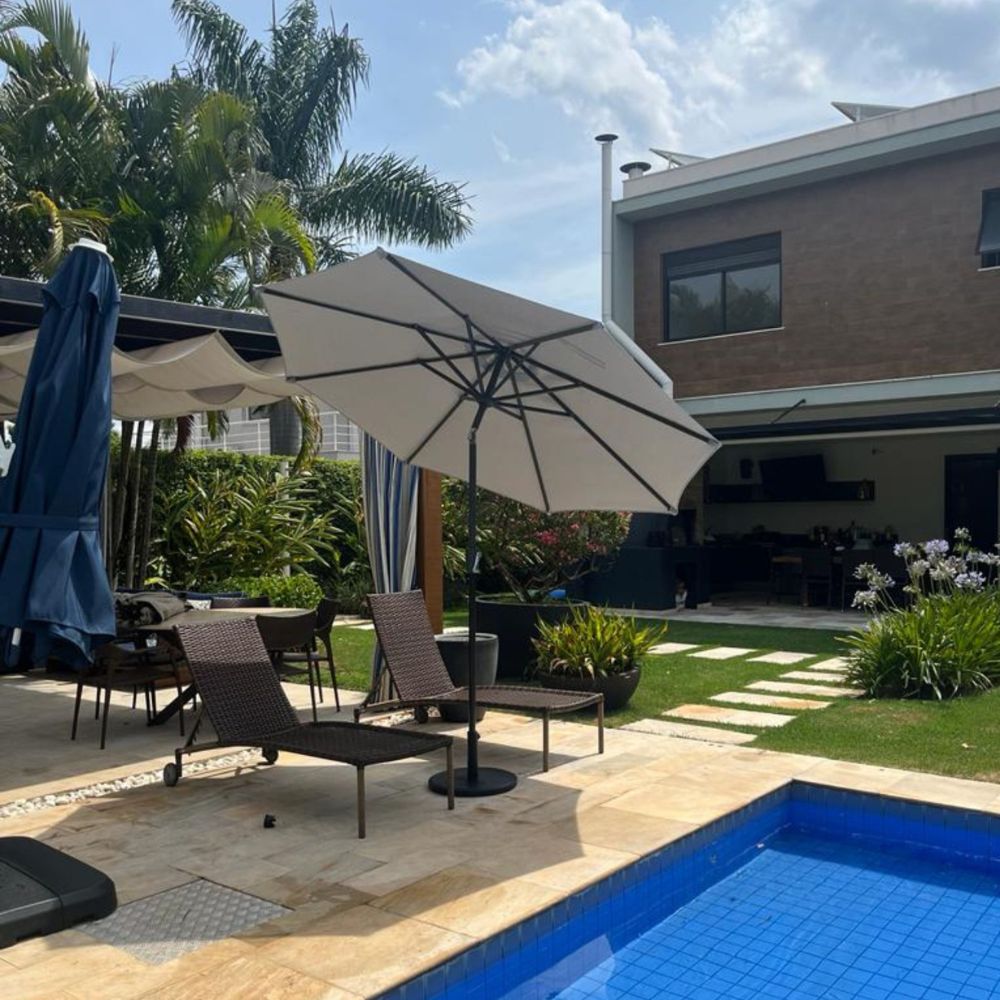 Ombrelone Santorini de centro, com cobertura bege, sombreando área de piscina e espreguiçadeiras de vime em ambiente externo.